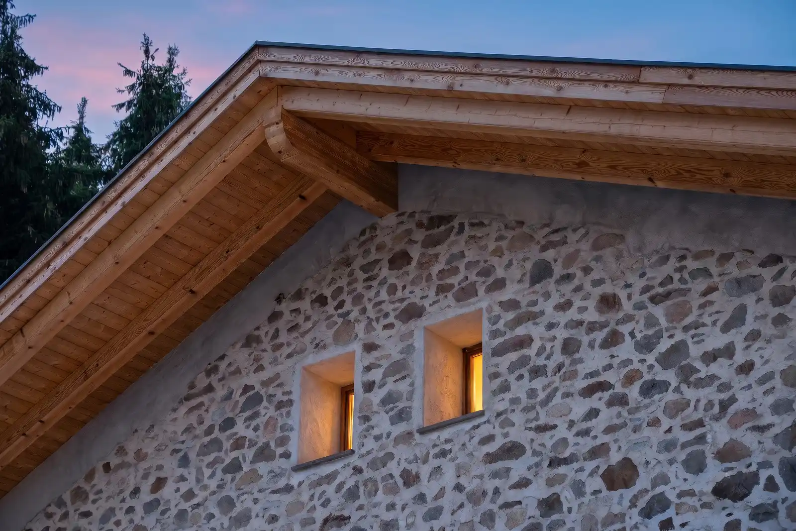 Due finestrelle di una casa in pietra all'ultimo piano, illuminate al tramonto, con dettaglio del tetto in legno ristrutturato