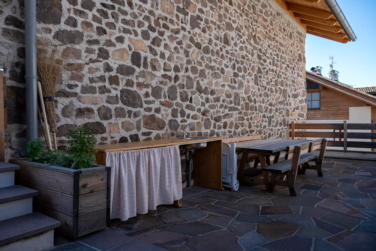 Terrazza con piastrelle, tavolo, panche e cucina esterna di una casa ristrutturata da 2R Group a Varena, in Val di Fiemme